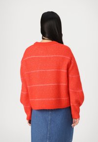 Punainen neulepaita, jossa on vaaleanpunaisia raitoja, pyöreä pääntie ja ylisuuria, laskostettuja olkapäitä. Paita on päällä farkkuhameen.