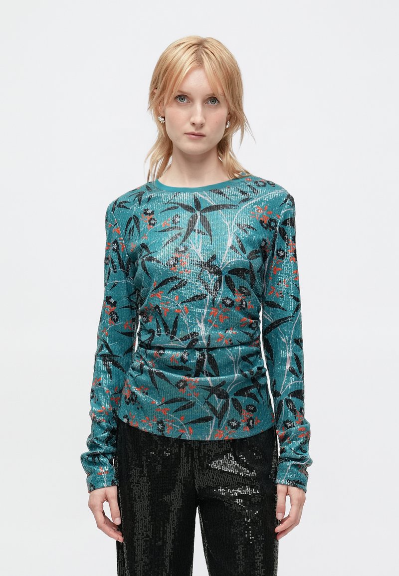 Teal langærmet bluse med blomsterprint i sort og rødt, lavet af et tekstureret stof. Sat sammen med sorte palietbukser.