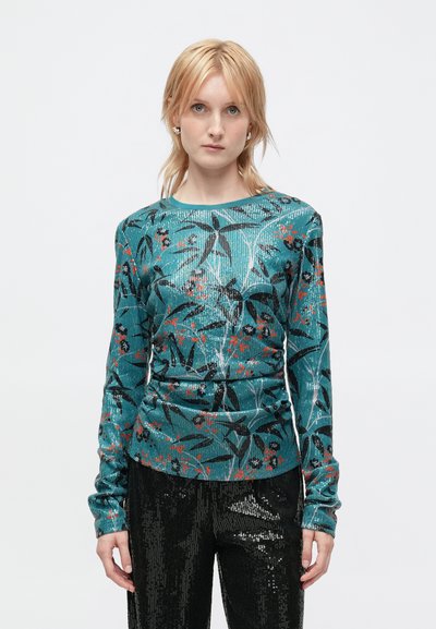 Haut à manches longues teal avec un motif floral en noir et rouge, fabriqué à partir d'un tissu texturé. Associé à un pantalon noir pailleté.