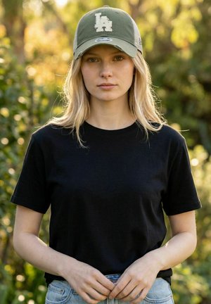 Young woman with long blonde hair wearing a green LA cap, black t-shirt, and blue jeans stands outdoors with hands clasped in front.
