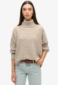 Suéter de punto beige con un cuello alto tipo mock, que presenta un patrón de zigzag texturizado. Lleva puesto unos jeans azul claro con un cinturón de cuero marrón.