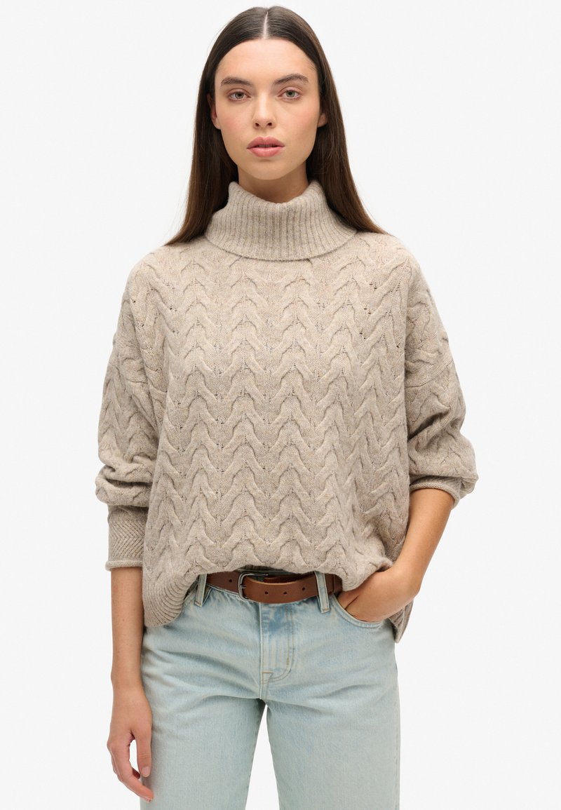 Suéter de punto beige con un cuello alto tipo mock, que presenta un patrón de zigzag texturizado. Lleva puesto unos jeans azul claro con un cinturón de cuero marrón.