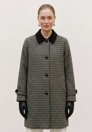 Femme portant un long manteau à motif pied-de-poule noir et gris, avec un col noir, des boucles d’oreilles en perles et des gants noirs, debout devant un fond uni.