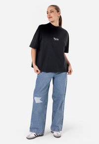 Juoda per didelė t-shirt su apvalia iškirpte ir balta logo, derinama su šviesiai mėlynomis suplyšusiomis pla�čiomis kelnėmis ir pilkais sportbačiais.