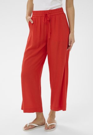 Kobieta ubrana w luźne czerwone spodnie ze sznurkiem i białe klapki, stojąca z jedną ręką w kieszeni na białym tle.