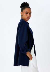 Blazer leggero di colore blu navy, con design aperto sul davanti, maniche a tre quarti e dettaglio con bottone ai polsini, abbinato a pantaloni bianchi.
