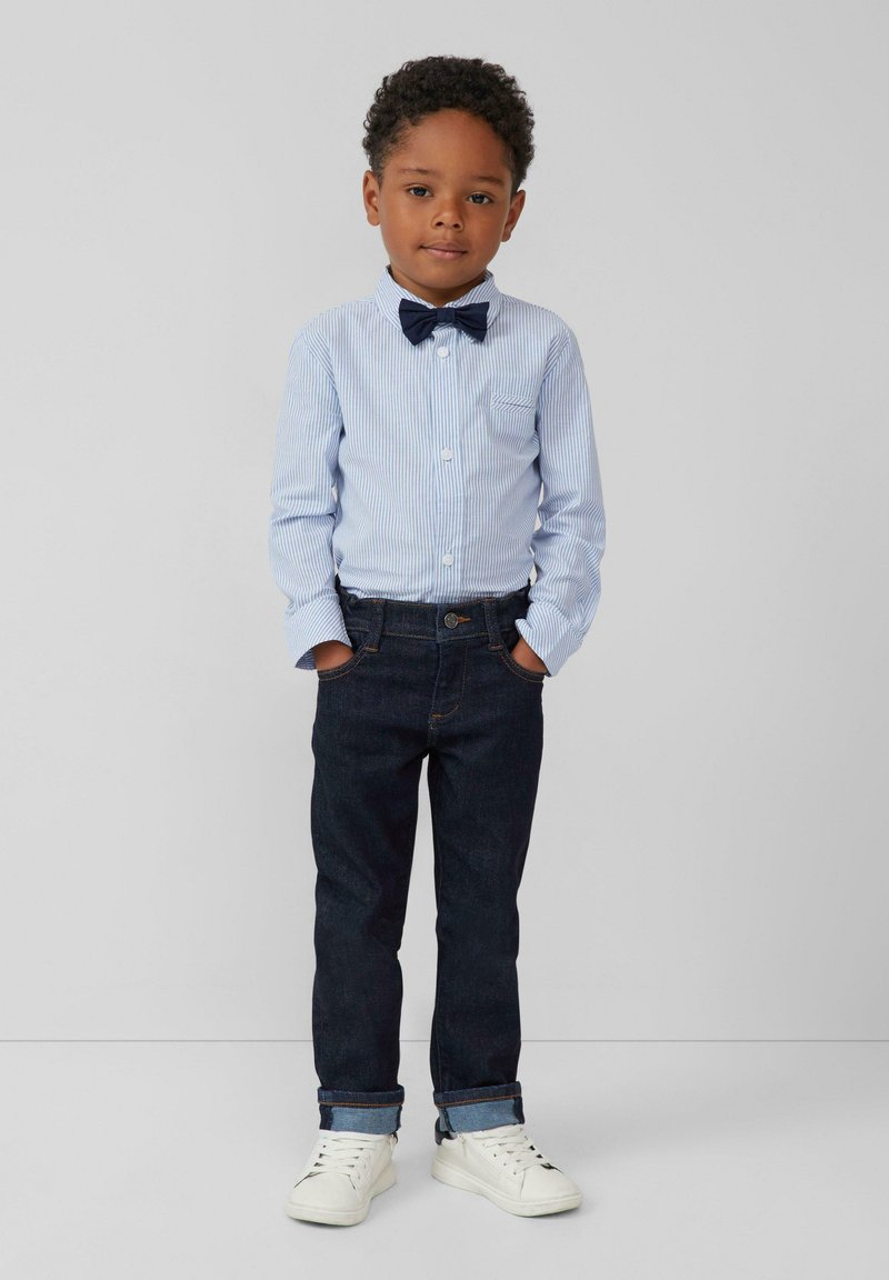 Blauw en wit gestreepte button-up shirt, donkere spijkerbroek en witte sneakers. Bevat een marineblauwe vlinderdas en opgerolde mouwen.