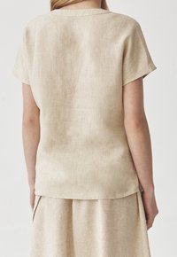 Žena oblečená v béžovém lněném topu s krátkými rukávy a odpovídající sukni, zobrazena zezadu na jednoduchém světlém pozadí.