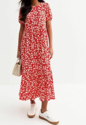 Kvinde iført en rød midi-kjole med hvidt blomsterprint, holder en beige vævet håndtaske og har hvide sneakers med gummisåler på.