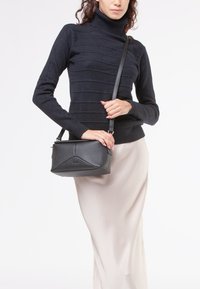 Borsa a tracolla nera in pelle con una forma strutturata, superficie testurizzata e pannello frontale geometrico, abbinata a un dolcevita nero aderente e a una gonna beige.