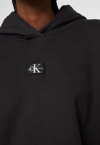 Melnas kokvilnas kapuces jaka ar priekšējo kabatu, kurai ir izšūts Calvin Klein Jeans logo uz krūts un kapuce ar aukliņu.