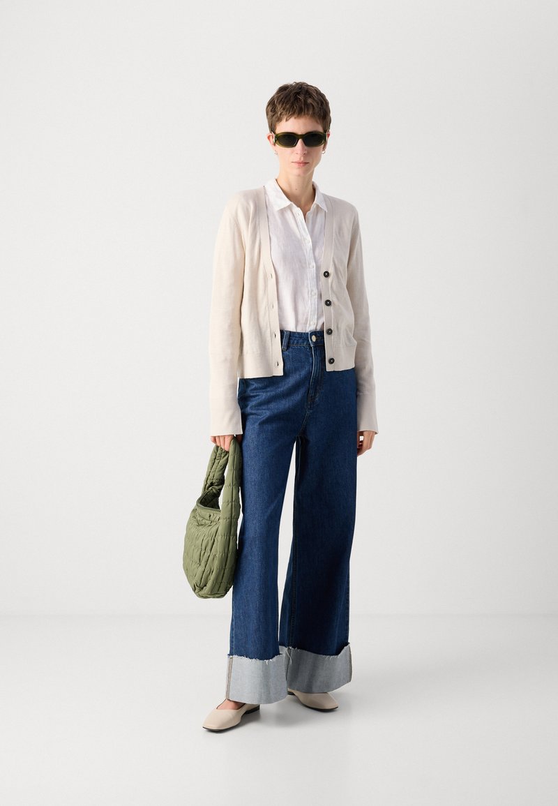 Weite blaue Denim-Jeans mit kontrastierenden grauen Säumen, weißem Hemd mit Knöpfen, cremefarbener Strickjacke, grüner puffy Tasche und Sonnenbrille.