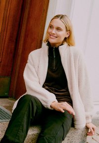 Heledest roosast fleecest kapuutsiga jakk, mis on kombineeritud tumepruuni korduroo ülemise osaga. Mustad püksid, tekstureeritud kanga ja mugava lõikega, millel on tõmblukkudetailid.