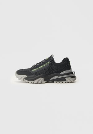Sneaker chunky nera e grigia con tessuto strutturato, suola spessa e testo verde "Versace Jeans Couture" sul lato.