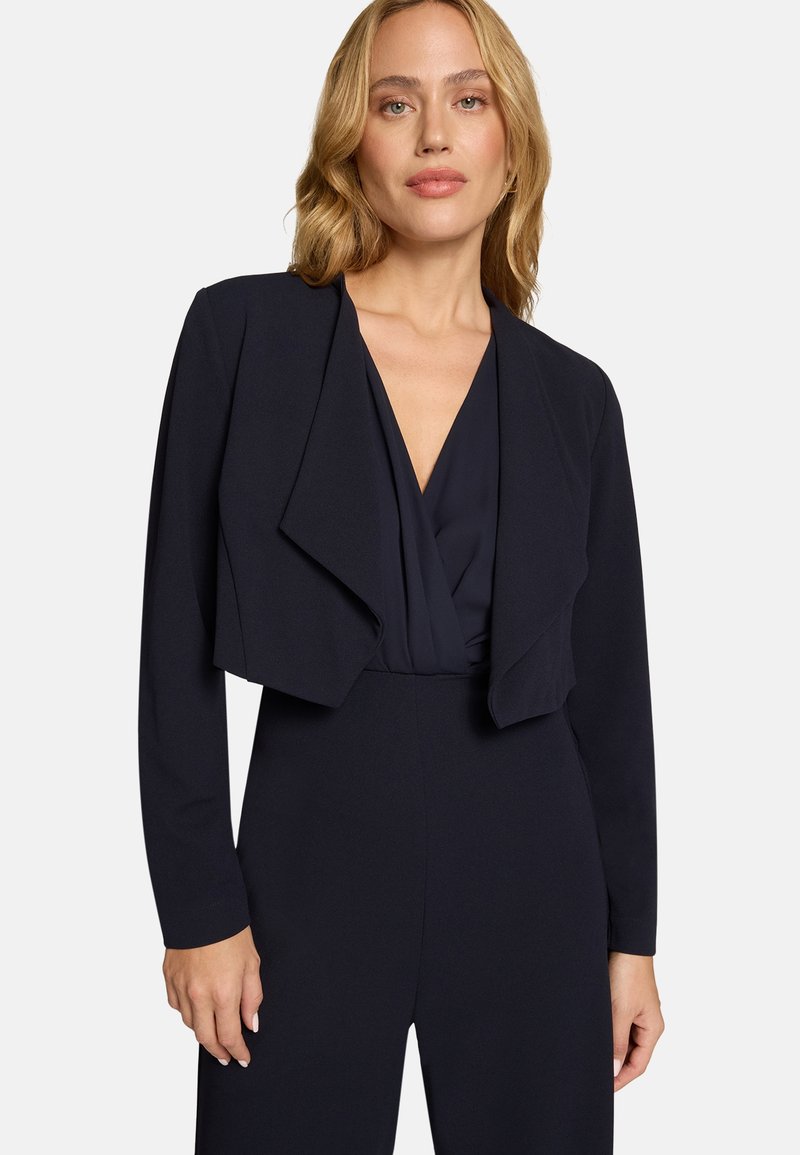 Femme blonde portant un blazer court bleu marine par-dessus une combinaison à décolleté en V profond, debout devant un fond clair uni.