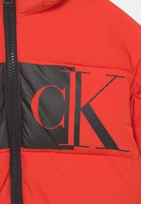 Rød og sort polstret jakke med sort lynlås og stort delvist synligt "CK" logo trykt på brystområdet.