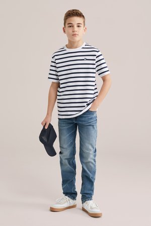 Fant, ki stoji in nosi belo majico z mornarsko modrimi črtami, modre kavbojke, bele superge ter v eni roki drži mornarsko modro kapo.