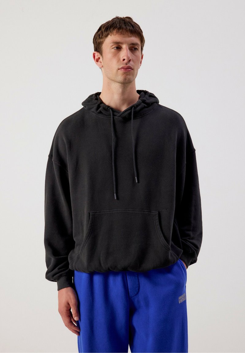Jeune homme aux cheveux courts et bruns, portant un sweat à capuche noir et un pantalon de survêtement bleu vif, debout sur un fond clair uni.
