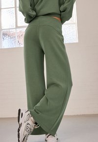 Green cotton wide-leg pants with a high waistband, featuring a smooth texture. Paired with white and gray sneakers. Bright window in background.