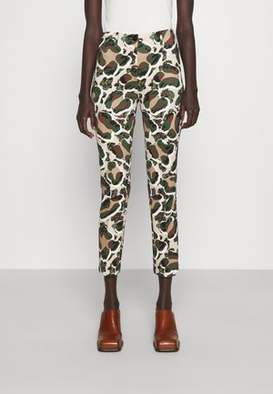 Dopasowane, skrócone spodnie z abstrakcyjnym nadrukiem kamuflażowym w odcieniach zieleni, brązu i beżu, noszone z brązowymi botkami ze kwadratowym noskiem na modelce o ciemnej karnacji.