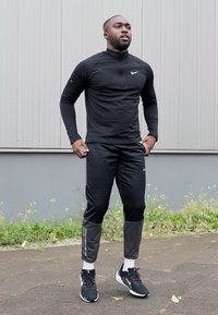 Crna Nike majica dugih rukava sa patentnim zatvaračem i rebrastom teksturom, uparena s crnim hlačama koje imaju sjajnu površinu i bijele detalje, te crnim sportskim cipelama.