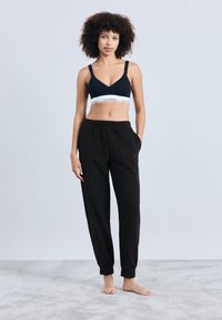 Juodas Calvin Klein sportinis liemenėlis su elastano juosta ir juodi sportiniai kelnės su elastinėmis rankovėmis, pagaminti iš minkštos medžiagos. Modelis stovi basomis.