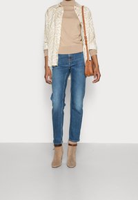 Camicia beige con motivo, maglione beige aderente, jeans blu e stivali alla caviglia color sabbia con accenti marroni. Piccola borsa a tracolla marrone.