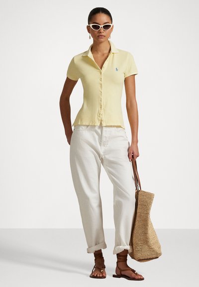 Dzeltena īsām piedurknēm polo krekls ar norādītu apkakli, pogu priekšu un mazāku zilu logotipu. Saskaņots ar baltām biksēm un austotu somu.