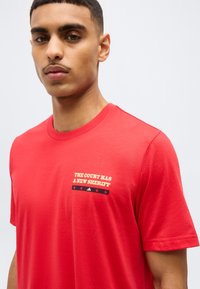 Νεαρός άνδρας που φορά κόκκινη μπλούζα με το κείμενο "THE COURT HAS A NEW SHERIFF" και γραφικά αστέρια στο στήθος, κοιτάζει μπροστά με ουδέτερη έκφραση.