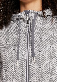 Sweat  à capuche gris zippé avec un motif en chevrons, doté d'une capuche doublée, de cordons de serrage et d'une fermeture éclair lisse. Tissu doux et texturé.