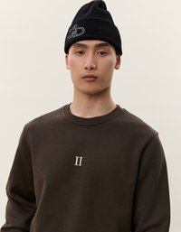 Felpa marrone con colletto rotondo e ricamo bianco "II" sul petto, abbinata a un cappellino di maglia nero con dettagli del logo bianchi.
