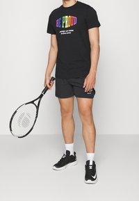 Czarny t-shirt z tekstem "BE PROUD" w wielokolorowym wydaniu, do tego szare szorty. Trzyma w ręku czarno-biały rakietę tenisową, ma na nogach czarne trampki.