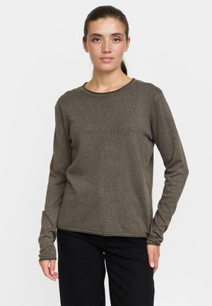 Jeune femme aux cheveux foncés attachés, portant un haut marron uni à manches longues et un pantalon noir, debout devant un fond blanc.