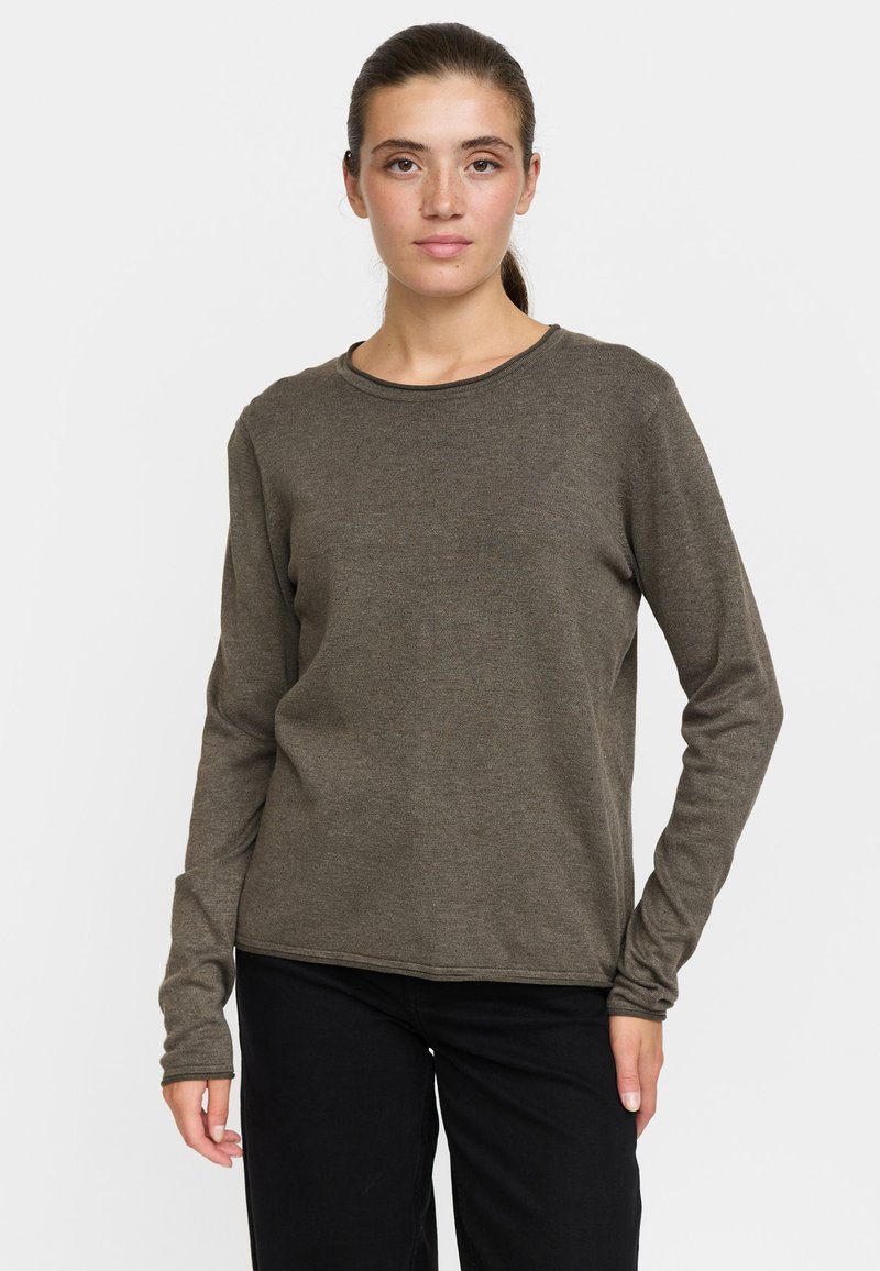 Jeune femme aux cheveux foncés attachés, portant un haut marron uni à manches longues et un pantalon noir, debout devant un fond blanc.