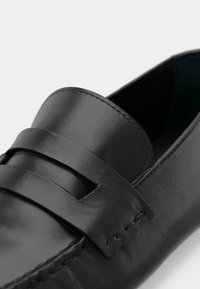 Μαύρα δερμάτινα μοκασίνια με λεία υφή, με φαρδιά λουράκι και ραφές κατά μήκος των άκρων. Εσωτερικός χώρος με ουδέτερο φόντο.