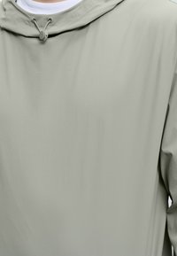 Ανοιχτό γκρι μπουφάν με κουκούλα και ρυθμιζόμενο κορδόνι, με λευκό γιακά ορατό από κάτω, εμφανίζεται από το στήθος και πάνω.