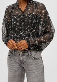 Person iført en gennemsigtig sort blomstret bluse med stofbeklædte knapper og puffede ærmer, parret med falmede grå jeans, hænderne foldet.