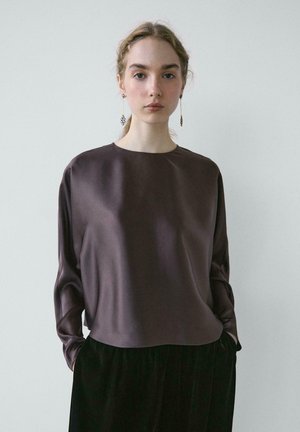 Jeune femme avec de longues boucles d'oreilles portant un chemisier ample en satin violet foncé et un pantalon noir, debout devant un fond clair uni.