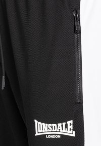 Fekete texturált anyag közeli képe fekete cipzárral és az alján fehér "LONSDALE LONDON" logóval nyomtatva.