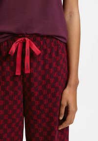 Burgunderrøde joggingbukser med rødt "HUGO"-logomønster, elastisk talje og bundet snøre. Blødt stof, afslappet pasform, synlig hånd.