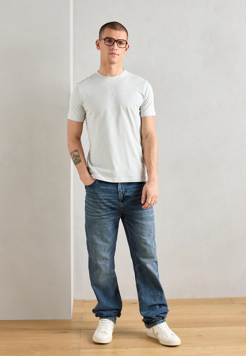 T-shirt à manches courtes gris clair, jeans bleus et sneakers blancs ; une personne se tient avec une posture détendue et porte des lunettes ; texture de tissu lisse.