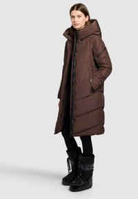 Pitkä, ruskea untuvatakki, jossa on korkea kaulus ja vetoketjusuljenta. Takissa on vaakasuoria tikattuja osioita ja sivutaskut. Yhdistetty mustiin talvisaappaisiin.