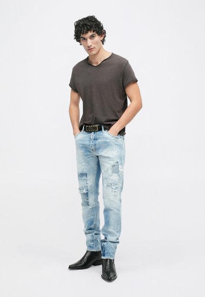 T-shirt gris foncé pour homme, jeans en denim bleu clair déchirés, ceinture noire avec des accents dorés, et bottes de cowboy noires à bout pointu.