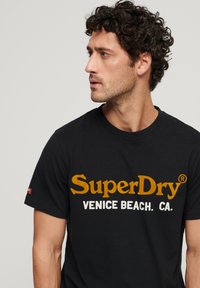 Czarny bawełniany t-shirt z dużym żółto-białym logo "SuperDry" i napisem "VENICE BEACH. CA." poniżej; okrągły dekolt, krótkie rękawy.