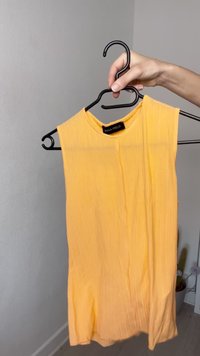 Top galben fără mâneci, cu guler rotund, confecționat din material plisat. Este așezat pe un umeraș din plastic negru; eticheta spune "ANNA FIELD."