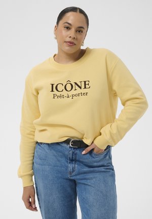 Kvinde iført en gul sweatshirt med teksten "ICÔNE Prêt-à-porter", blå jeans, sort bælte, hånd i lommen og gyldne hoops øreringe.