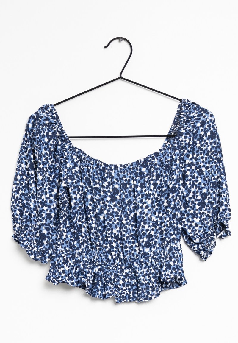 Blouse florale bleue avec des manches bouffantes, un col froncé et un ourlet volanté. Fabriquée en tissu léger avec un motif texturé dans des nuances de bleu variées.