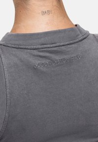 Grijze mouwloze shirt met een ronde halslijn en subtiele stiksels. "GOODFOR NOTHING" is in een donkere tint borduurwerk op de achterkant.