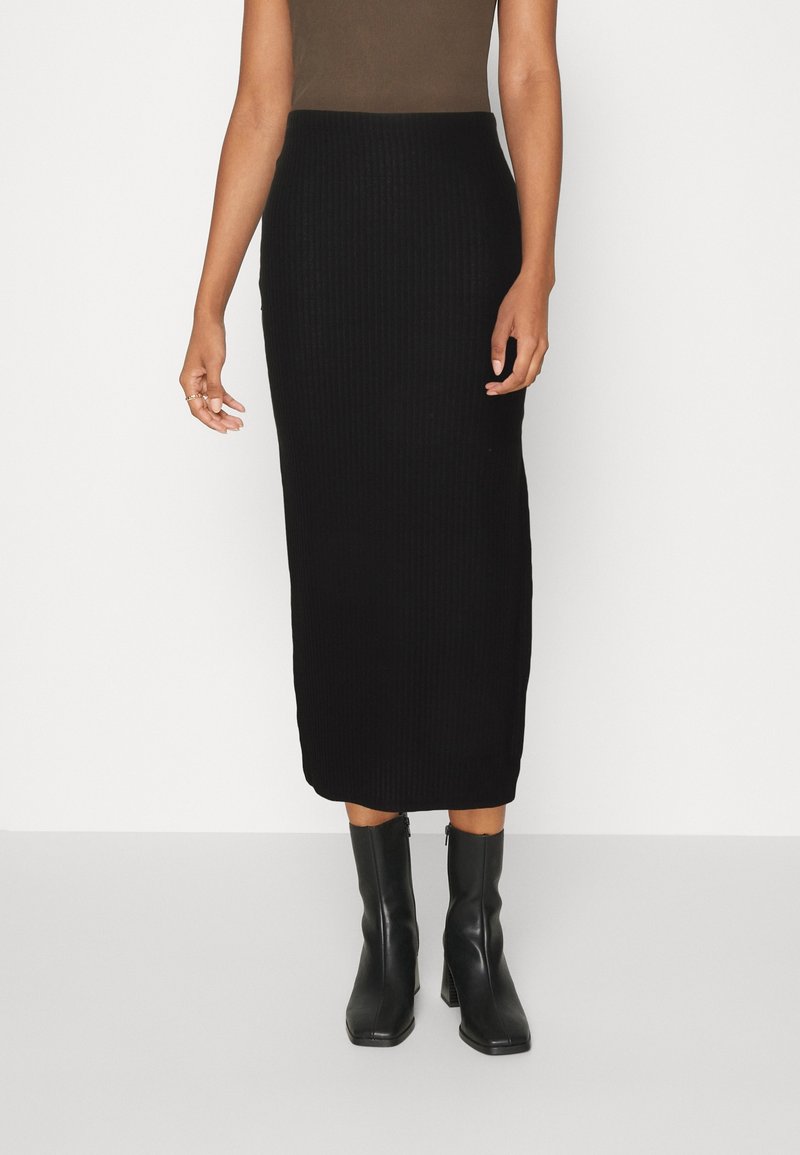 Femme portant une jupe midi noire moulante côtelée, un top marron sans manches et des bottines noires à talons, debout devant un fond blanc.