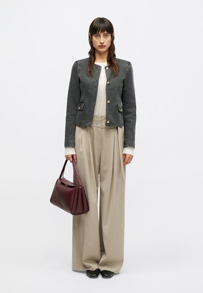 Žena v tmavě šedém kabátu na knoflíky, béžových širokých kalhotách, s burgundskou kabelkou, stojící na jednobarevném pozadí.